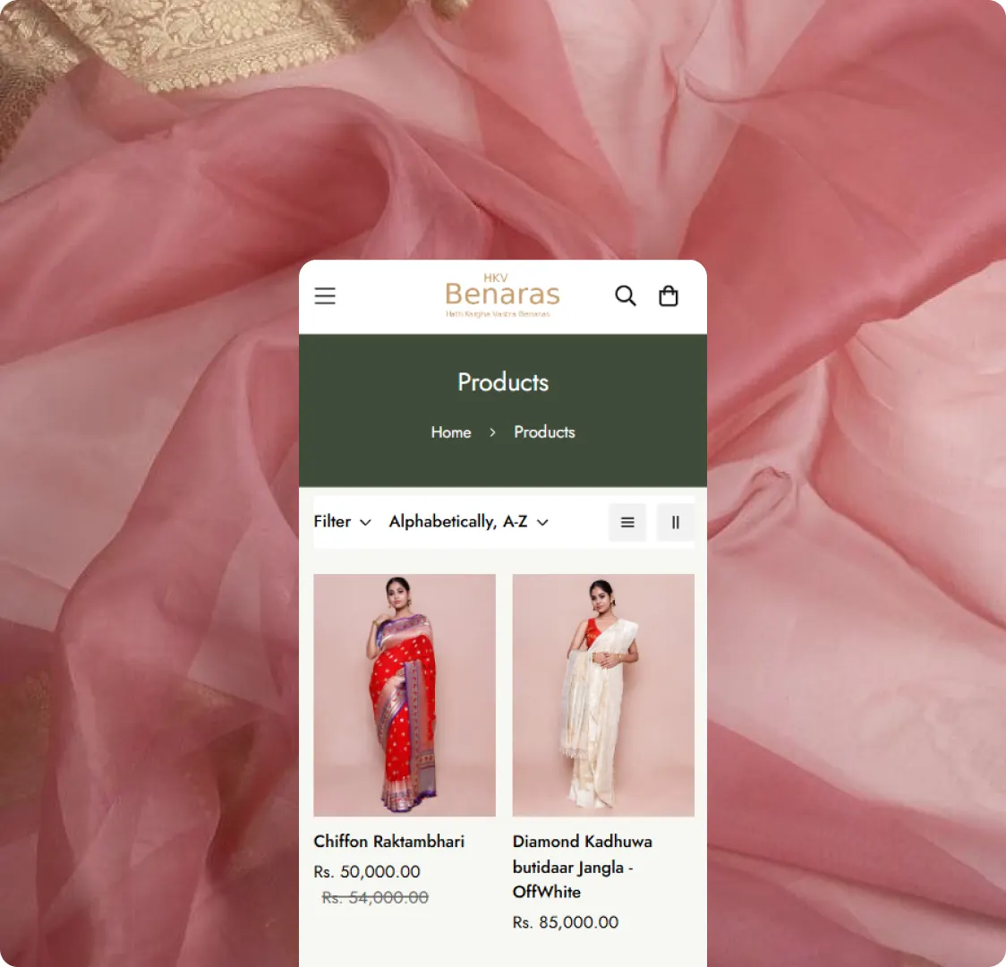 A phone on a pink fabric background showing the HKV Benaras Products listing page, with filter and sort controls above two product cards.