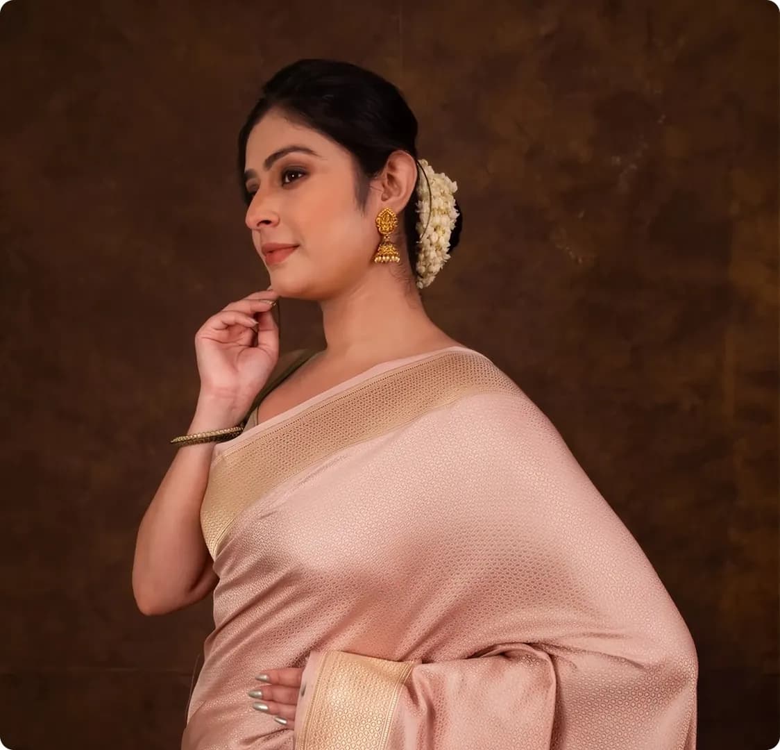 A photograph of a woman in a pale pink silk saree against a dark brown backdrop.
